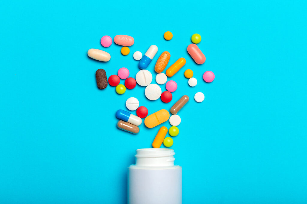 Pills and tablets of various colors are falling from a white bottle onto a blue surface.